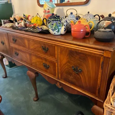 Vintage Queen Anne style wooden buffet with cabriole legs, featuring a collection of decorative teapots and figurines on top.