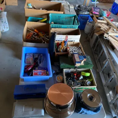 Folding table filled with various garage and household items, including cookware, hand tools, hardware, and plastic storage crates.