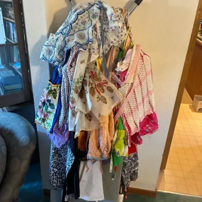 Collection of vintage kitchen aprons and linens featuring various floral, pinecone, and crocheted patterns hung over a stand.