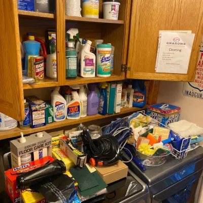 An assortment of household cleaning supplies, sponges, trash bags, and laundry items displayed inside a wooden cabinet and atop appliances.