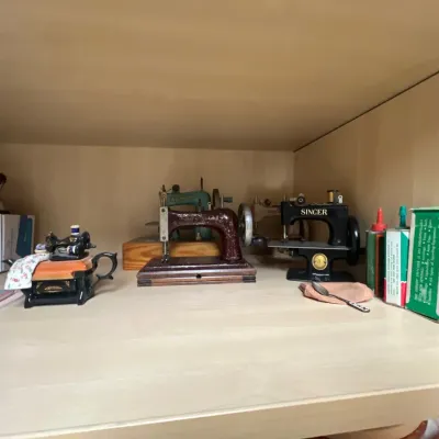 Collection of vintage toy sewing machines, including a black Singer model and a maroon cast iron model, displayed on a wooden shelf...