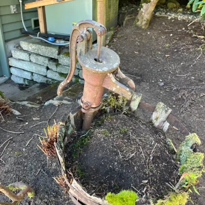 Vintage cast iron hand water pump mounted inside a weathered, soil-filled wooden barrel planter.