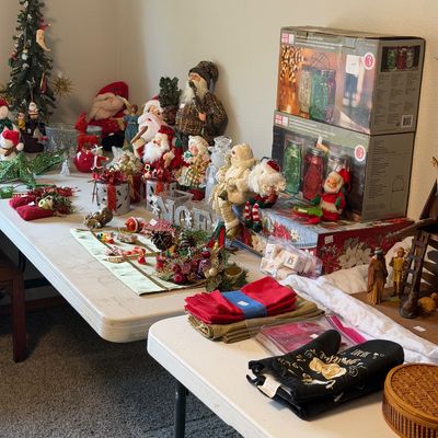 Sale Photo Thumbnail #32: Assortment of Christmas decorations, including Santa figurines, a small artificial tree, holiday linens, and boxed decorative jars, displayed on folding tables.