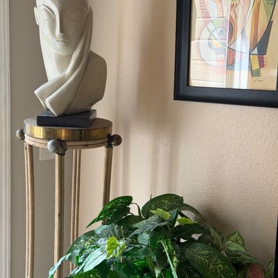 Sale Photo Thumbnail #17: Art Deco style ceramic bust of a woman with a draped headpiece, displayed on a tall, slender pedestal stand alongside a framed abstract print.