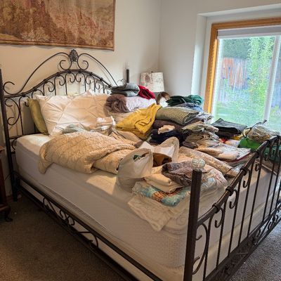 Sale Photo Thumbnail #29: Wrought iron queen-size bed frame with scrollwork details, shown piled with various blankets, quilts, and linens for sale.