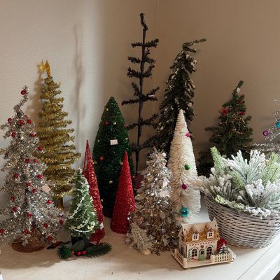Sale Photo Thumbnail #33: Collection of vintage and modern tabletop Christmas trees in various styles, including tinsel, bottle brush, and flocked designs, displayed alongside a small decorative house.