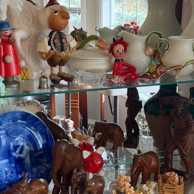 Sale Photo Thumbnail #8: A glass display cabinet filled with various collectibles, including hand-carved wooden elephants, ceramic figurines, a vintage red devil doll, and decorative white pitchers.