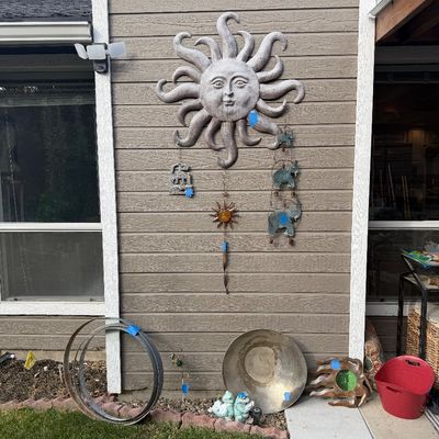 Sale Photo Thumbnail #39: Large cast stone sun face wall plaque, accompanied by hanging wind chimes, metal garden hoops, and various outdoor decor items.