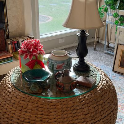 Sale Photo Thumbnail #20: Woven wicker or seagrass pouf coffee table with a glass top, styled with a black table lamp, decorative ceramics, and a floral arrangement.