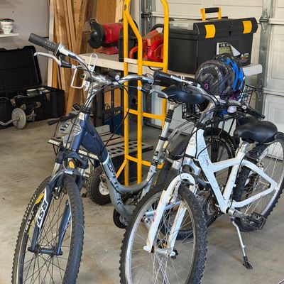 Sale Photo Thumbnail #35: Two Trek hybrid bicycles, including a blue Trek 7300 Multitrack and a white Trek Navigator, shown with helmets and garage tools in the background.