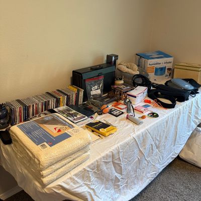 Sale Photo Thumbnail #27: Assorted household items displayed on a table, including a stack of rug pads, a row of CDs, a humidifier in its box, and various small electronics and accessories.