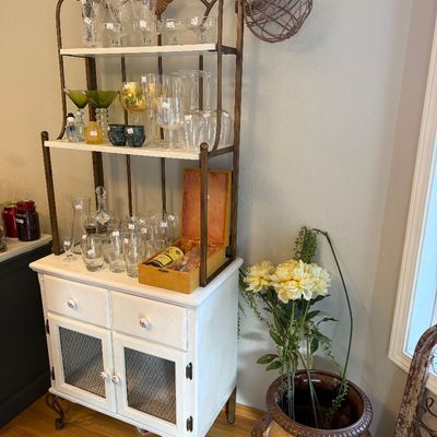 Sale Photo Thumbnail #11: Vintage white-painted baker's rack with a brass-finished metal frame, featuring two upper shelves displaying glassware and a lower cabinet with wire-mesh doors.