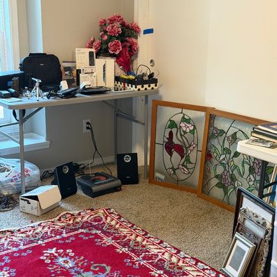 Sale Photo Thumbnail #26: Assorted household items including a red patterned area rug, two framed stained glass window panels, small electronics, and a folding table with various accessories.