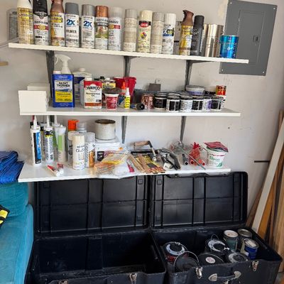 Sale Photo Thumbnail #36: Assortment of spray paints, paint thinners, sealants, and various home improvement supplies organized on wall-mounted shelves, with two large black plastic storage trunks containing paint cans below.