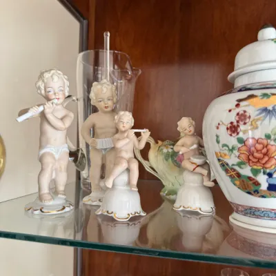 Sale Photo Thumbnail #131: Collection of vintage porcelain cherub figurines playing musical instruments, displayed on a glass shelf alongside a floral ginger jar and glassware.