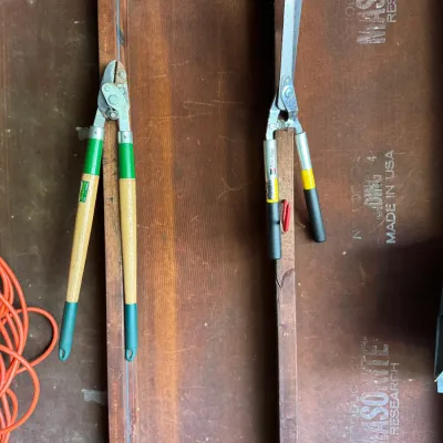 Two pairs of vintage garden shears, including a pair of wooden-handled loppers and a pair of metal hedge shears, resting on a Masonite...