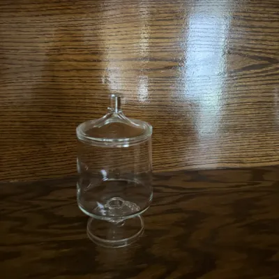 Sale Photo Thumbnail #143: Small clear glass apothecary jar with a matching lid, displayed on a dark wood surface.