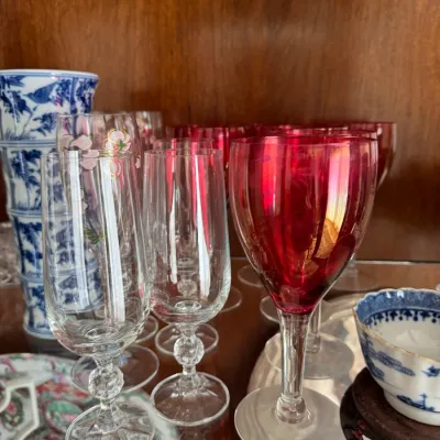 Sale Photo Thumbnail #133: Assorted vintage glassware including clear champagne flutes with decorative stems and ruby red wine goblets, displayed alongside blue and white porcelain pieces.