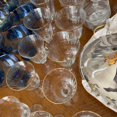 Sale Photo Thumbnail #107: Collection of vintage glassware, featuring gold-rimmed ribbed wine glasses, blue goblets, and cut crystal bowls on a wooden table.