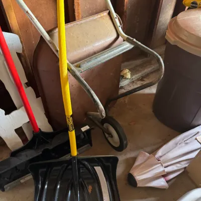 Steel Core snow shovel with a yellow handle, shown alongside a weathered metal wheelbarrow and other outdoor utility items.