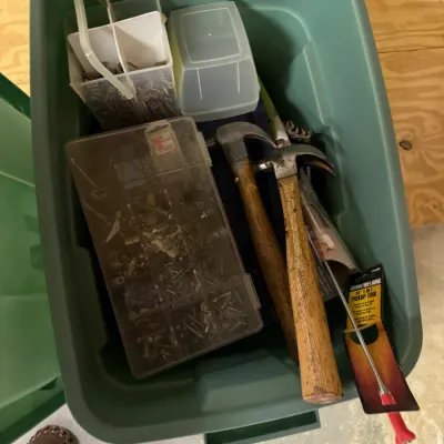 Green plastic storage bin containing various hand tools and hardware, including two wooden-handled claw hammers, a plastic organizer...