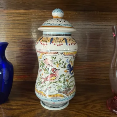 Hand-painted Italian majolica ceramic lidded jar, featuring a colorful bird and floral motif on a wooden shelf.
