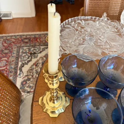 Sale Photo Thumbnail #111: Pair of vintage brass candlesticks with scalloped bases, shown with cream taper candles alongside blue glassware and a cut glass cake stand.