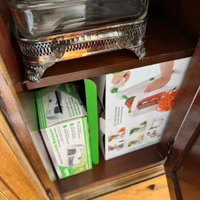 Sale Photo Thumbnail #104: Vintage glass baking dish in a silver-plated footed holder, shown stored in a cabinet above boxed kitchen gadgets including a knife sharpener and a spiralizer.