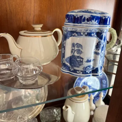 Sale Photo Thumbnail #136: Vintage blue and white porcelain lidded jar with a willow pattern, displayed alongside a cream-colored teapot with gold trim and various glassware on glass shelves.