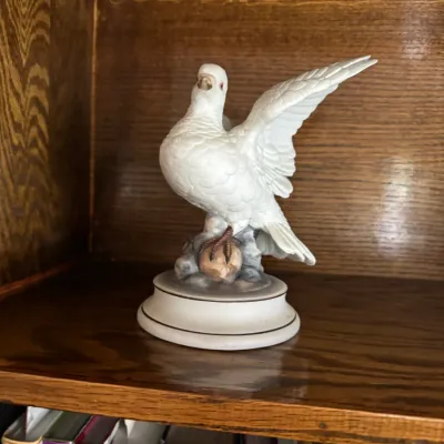 Sale Photo Thumbnail #183: Vintage white porcelain dove figurine with outstretched wings, perched on a rocky base and displayed on a wooden bookshelf.