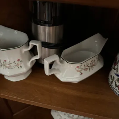 Vintage porcelain gravy boat and creamer set featuring a delicate pink floral and ribbon motif with green trim, displayed on a wooden shelf.