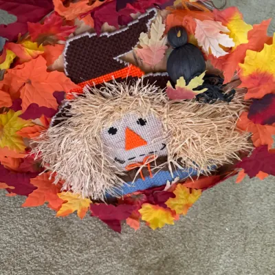 Sale Photo Thumbnail #174: Handcrafted autumn wreath featuring a needlepoint scarecrow face with raffia hair, surrounded by colorful faux maple leaves.