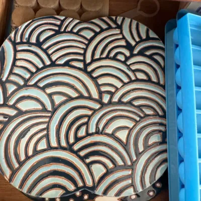 Ceramic trivet featuring a raised, stylized wave pattern in light blue and dark brown glazes, stored in a drawer.