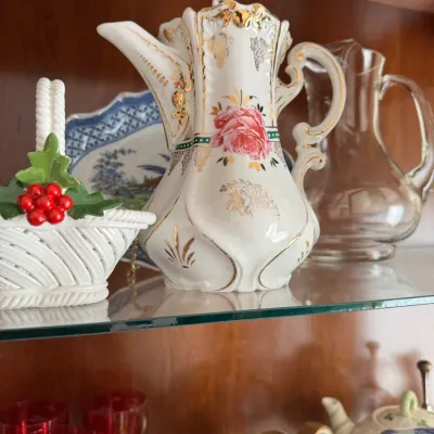 Sale Photo Thumbnail #135: Vintage porcelain coffee pot featuring a pink rose motif and ornate gold-gilded accents, displayed alongside a ceramic holly basket and a clear glass pitcher on a glass cabinet shelf.