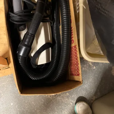 Oreck handheld canister vacuum cleaner with hose and attachments, stored in its original cardboard box.