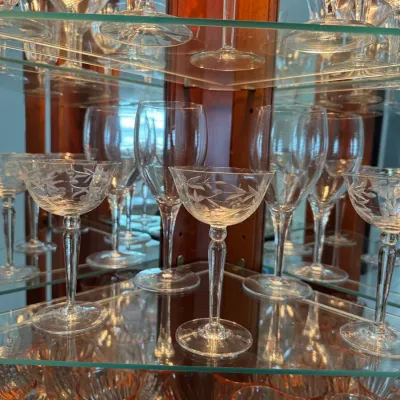 Sale Photo Thumbnail #118: Collection of vintage crystal stemware, including etched coupe glasses and cut-glass goblets, displayed on glass shelves in a wooden china cabinet.