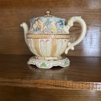 Sale Photo Thumbnail #147: Vintage Capodimonte-style porcelain teapot with matching warming stand, featuring a relief design of cherubs, gold accents, and an ornate handle.