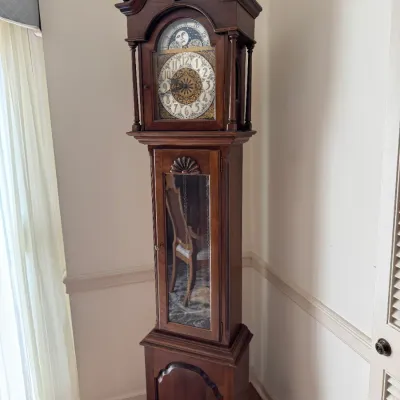 Sale Photo Thumbnail #114: Vintage mahogany grandfather clock featuring a swan neck pediment with finials, an ornate dial with a moon phase indicator, and a glass-front pendulum door.