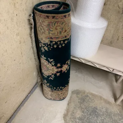 Rolled Persian-style area rug featuring a deep teal field with traditional floral motifs and a beige patterned border.