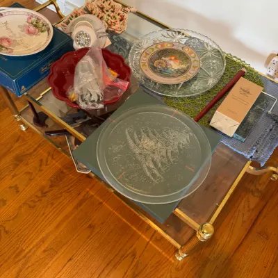 Sale Photo Thumbnail #159: Assorted decorative plates, glassware, and holiday tabletop items displayed on a two-tier brass and glass bar cart.
