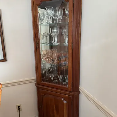 Sale Photo Thumbnail #117: Traditional cherry wood corner hutch featuring a glass-front display cabinet with glass shelves, filled with crystal stemware, above a lower wooden storage cabinet.