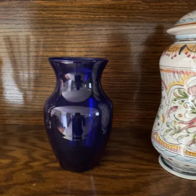Sale Photo Thumbnail #194: Cobalt blue glass vase with a flared rim, displayed on a wooden shelf next to a hand-painted ceramic jar.