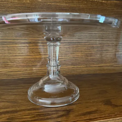 Sale Photo Thumbnail #169: Vintage clear glass pedestal cake stand with a simple, elegant design, displayed on a wooden shelf.