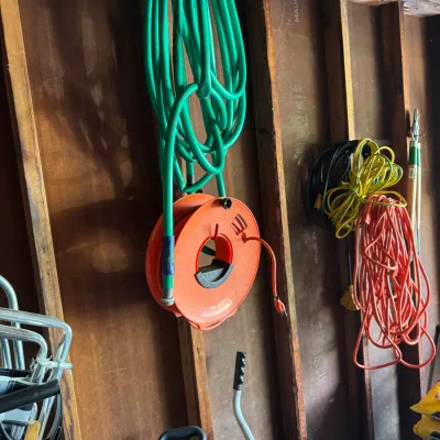 Assorted garden and utility equipment hanging on a wooden wall, including a green garden hose, an orange extension cord reel, and...