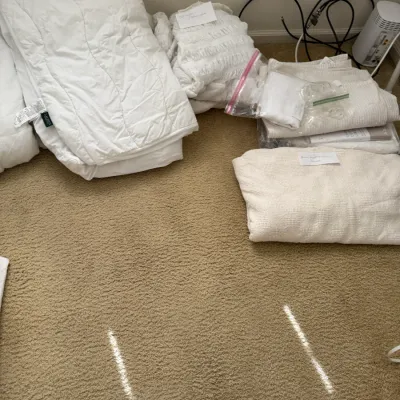 Assorted white and cream bedding, including comforters, blankets, and sheets, arranged on a carpeted floor.