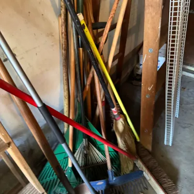 Assorted long-handled lawn and garden tools, including rakes, brooms, and shovels, leaning against a garage wall.