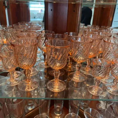 Sale Photo Thumbnail #119: Collection of vintage pink depression glass stemware featuring a swirl pattern, displayed on glass shelves in a wooden cabinet.