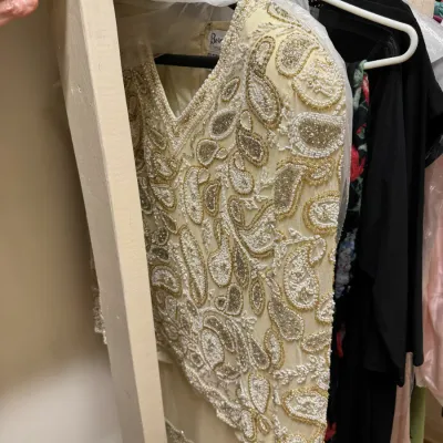 Vintage cream-colored long-sleeve blouse featuring intricate gold and white beadwork in a paisley pattern, hanging in a closet.