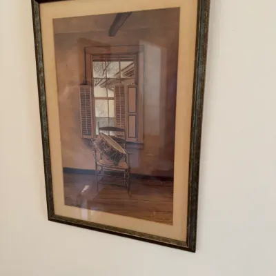 Sale Photo Thumbnail #157: Framed art print depicting a rustic interior scene with a drum resting on a chair by a window, presented in a dark, textured frame with a tan mat.