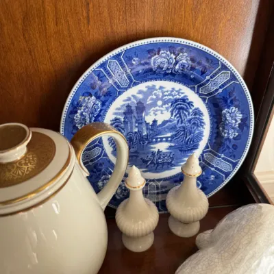 Vintage blue and white transferware plate featuring a scenic landscape, displayed alongside a white and gold porcelain teapot and...